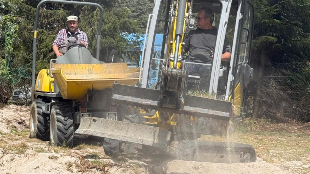 Ein Bagger schaufelt Sand in einen Lader um den Beachvolleyplatz zu reinigen.